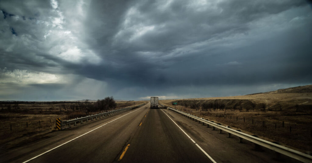 A truck driving on an empty road with storm clouds in the sky overhead.