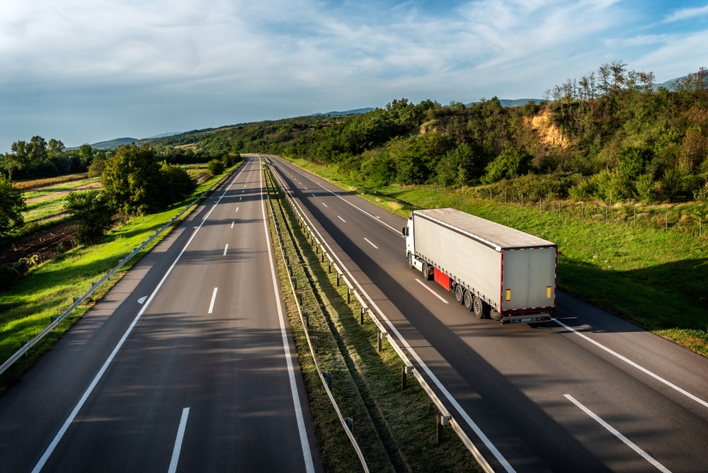 A truck driving along a highway with greenery and hills surrounding it.